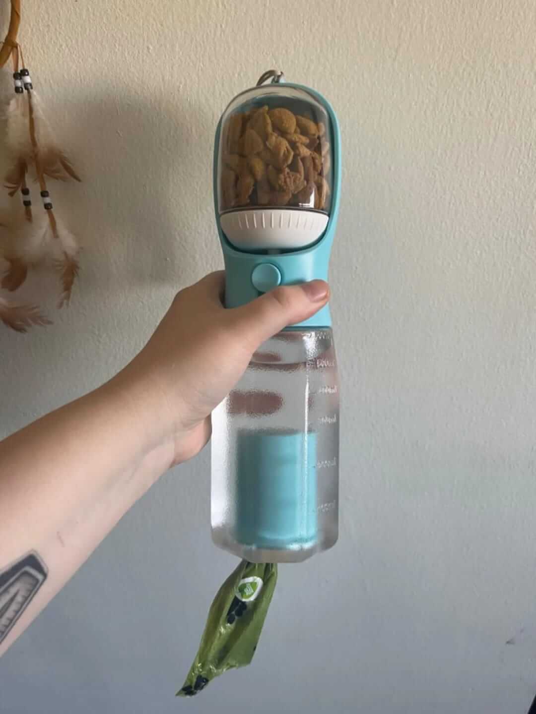 Hand holding a blue pet water bottle with treats inside against a plain wall.